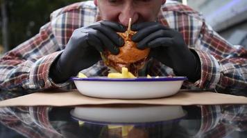 A man in a plaid shirt and black gloves taking a bite of a juicy burger and chew. Sauce is dripping from the hamburger held by the man over plate of french fries sitting at a table outdoor - close up. video