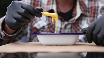 A man in a plaid shirt and black gloves dunks French fries into a bowl of melted cheese. The sauce is dripping from the potato held by the man sitting at a table outdoor - close up. video