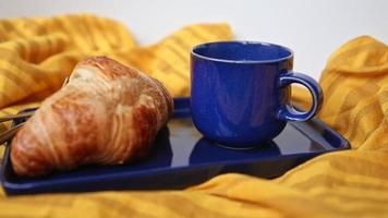 Taza azul humeante y plato con croissant horneado francés caliente sobre un paño naranja, preparado para un cliente habitual en la cafetería y la panadería. desayuno o pausa para el café en la cafetería. no hay gente en el video. video