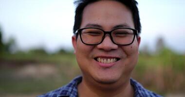 Portrait Close up of an Asian young happy farmer man with glasses looking at camera and smiling with a farmland background. Agriculture farming concept. Slow motion. video