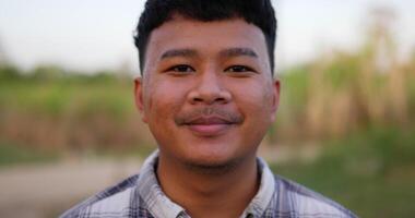 retrato próximo de um jovem fazendeiro feliz asiático olhando para a câmera e sorrindo com um fundo de terras agrícolas. conceito de agricultura agrícola. câmera lenta. video