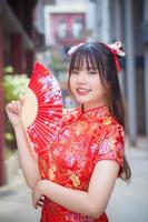 Cute Asian young woman in red cheongsam dress in Chinese new year theme stands smiling happily looking at the camera holds fan among old city center in Chinese new year theme. photo