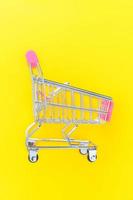 Small supermarket grocery push cart for shopping toy with wheels isolated on yellow colourful trendy modern fashion background. Sale buy mall market shop consumer concept. Copy space.