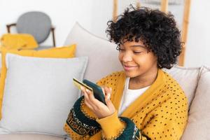 African american woman shopping online holding smartphone paying with gold credit card. Girl sitting at home buying on Internet enter credit card details. Online shopping ecommerce delivery service.