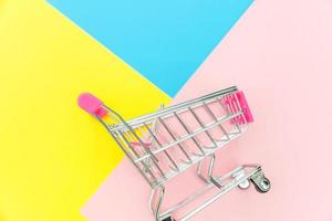 Small supermarket grocery push cart for shopping toy with wheels isolated on blue yellow pink pastel colorful trendy geometric background Copy space. Sale buy mall market shop consumer concept. photo