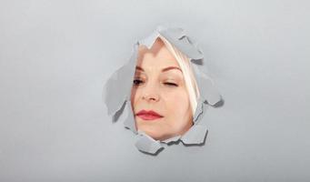 Surprised woman looking playfully in torn paper hole, has excited cheerful expression, looks through breakthrough of gray background. Wow, great news photo