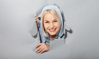 Charismatic joyful woman with blonde hairstyle, looking playfully, wears fashionable hoody, stands in torn paper hole, notices funny pleasant. Gray background. photo