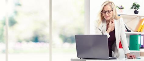 Beautiful middle aged woman using laptop, standing in office near the window. photo