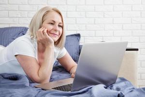 Smiling attractive woman lying on bed using laptop communicating online at home, happy girl typing on computer, enjoying internet shopping or chatting in social networks in bedroom. photo