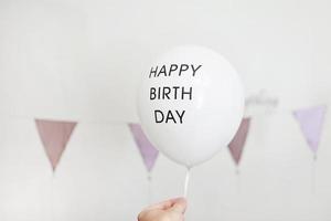 Unrecognised hand holding a white balloon with black inscription Happy birthday in decoration for the holiday. colourful garland flags at wall on background photo