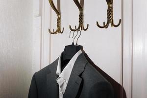 Man's dark grey suit jacket and white shirt hanging on gold metal hook on white wall background. photo