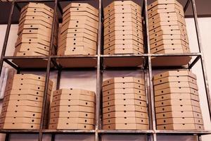 servicio de entrega de alimentos. pilas altas de cajas de pizza planas de cartón marrón en una estantería de metal listas para su entrega. cajas de embalaje artesanales con pizza en stock en almacenamiento foto