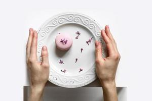 cropped view of woman holding plate with pink delicious French macaroon or macaron with lilac flowers on white background photo