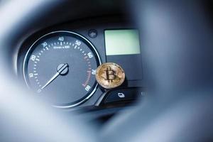 The physical coin is a gold bitcoin on the dashboard of the car next to the fuel consumption photo