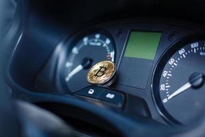 The physical coin is a gold bitcoin on the dashboard of the car next to the fuel consumption photo