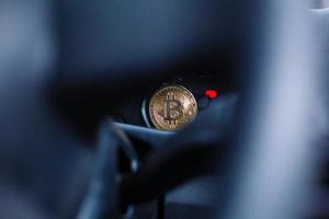 The physical coin is a gold bitcoin on the dashboard of the car next to the fuel consumption photo