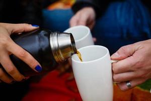 Young couple on a picnic in a park man pouring hot coffee from a thermos bottle focus is on the photo