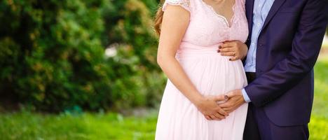 Pregnant girl and her husband are happy to hug, hold hands, round stomach, stand on grass in the outdoor in the garden background with trees. Close up. full length. Looking down at the stomach photo