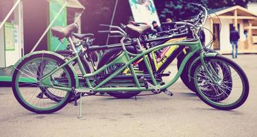 A green colored tandem bicycle with scarlet wheels is parked in a car park near the bike path photo