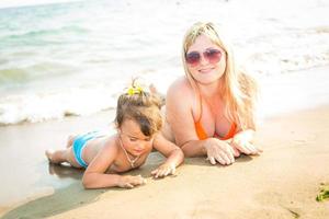 Portrait of beautiful young parents and cute little girl in swimsuits and sun glasses smiling, lying on the sunny beach. Parents are looking at their daughter photo