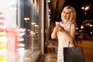 hermosa joven mujer de moda con bolsas de compras cerca de la ventana de la tienda escribe un teléfono inteligente sms, en la calle de la ciudad de noche foto