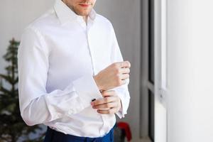Man buttons up his white shirt standing in the front of a bright window photo