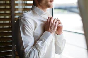 Man buttons up his white shirt standing in the front of a bright window photo