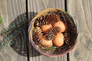 Easter eggs in a basket of 4 with pine leaves and pine cones on a light wooden background. photo