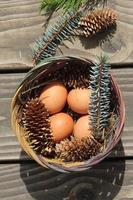 Easter eggs in a basket of 4 with pine leaves and pine cones on a light wooden background. photo
