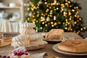 White airy meringue cake, honey cake and pie on the festive table, against the backdrop of a Christmas tree with a garland