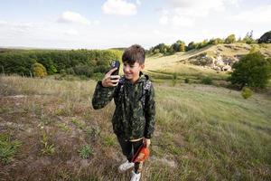 Activity on sunny autumn day, boy exploring nature. Kid wear backpack hiking and making photo on phone.