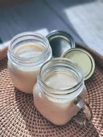 2 dos vasos de tarros de albañil con yogur casero natural, yogur, en la bandeja de mimbre tejida a mano, cesta de mimbre, vista en ángulo alto. color de tema suave estilo café coreano, vertical foto
