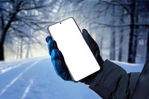 teléfono móvil en mano con guante de invierno. árboles cubiertos de nieve en el fondo. pantalla aislada para promoción de aplicaciones foto