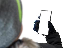 un tipo con una chaqueta con gorra y guantes sostiene un teléfono móvil. el fondo y la pantalla del teléfono están aislados en blanco para una maqueta, presentación del diseño de una aplicación o sitio web
