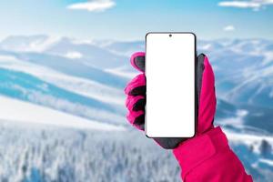 utilizando un teléfono móvil en la estación de esquí. pantalla aislada para promocionar la aplicación de esquí o montañismo. teléfono en mano con guante rosa. estación de esquí en segundo plano