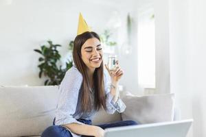 copie la foto espacial de una joven alegre que tiene un evento de celebración de cumpleaños con un amigo a través de una videollamada. ella está haciendo un brindis de celebración con una copa de vino blanco hacia la cámara del portátil.