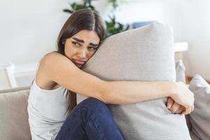 mujer joven abrazando la almohada con depresión y llorando en el sofá en casa. dama milenaria solitaria que se siente estresada y desesperanzada, sufre de trastornos del estado de ánimo o tiene problemas psicológicos