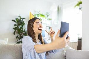 Virtual Party. Happy young woman in hat having video conference online meeting with friends and family, holding glass of wine, toasting and celebrating birthday, staying at home photo