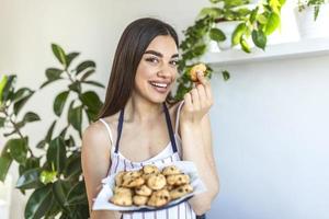 joven ama de casa hermosa sosteniendo galletas recién horneadas en una bandeja en la cocina. foto