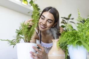 Woman caring for plants next to steam aroma oil diffuser on the table at home, steam from humidifier. Humidification of air in apartment during the period of self-isolation due to coronavirus pandemic photo