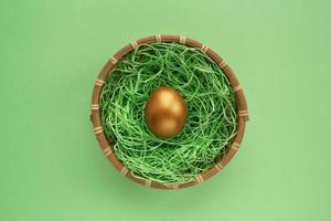 Golden egg in wooden rattan wicker basket on green background. Financial investment business concept. Easter egg hunt festive design composition. Top view, flat lay. World egg day. Close-up photo