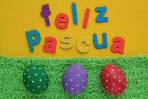 feliz pascua, letras felices de pascua en español sobre fondo amarillo y huevos de color sobre hierba verde. banner de vacaciones, composición creativa flatlay. tarjeta de felicitación, afiche, pancarta. vista superior concepto festivo foto