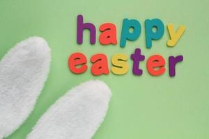 Happy Easter colored lettering and white bunny fur ears on green background. Holiday spring composition creative concept. Idea for funny event postcard, banner, poster, party. Top view, flatlay photo