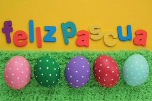 Feliz Pascua, Happy Easter letters on spanish on yellow background and color eggs on green grass. Holiday banner, flatlay creative composition. Greeting card, poster, banner. Top view festive concept photo