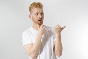 Happy young redhead guy, red stylish beard isolated. Man pointing copy space for advertising. Male in white t shirt, trend haircut. Successful young entrepreneur. Surprised shocked excited man face photo