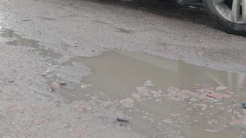 baches llenos de agua en una carretera asfaltada con coches que pasan. clima lluvioso. el coche conduce a través de charcos en carreteras asfaltadas rotas después de la lluvia durante el día. gotas de lluvia y coches en la carretera. video