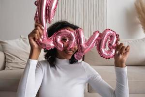 A beautiful young African-American woman with balloon in the shape of the inscription love sits near sofa. Woman on Valentine's Day. Symbol of love photo
