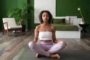 joven mujer afroamericana sana y tranquila en ropa deportiva sentada en casa haciendo yoga, meditando y respirando por la mañana. concepto de salud mental foto