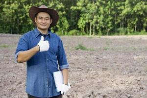 Asian man agronomist wears hat, blue shirt, holds paper clipboard, thumbs up, stand at agriculture land. Concept, Agriculture research , survey and study to develop soil quality for growing crops. photo