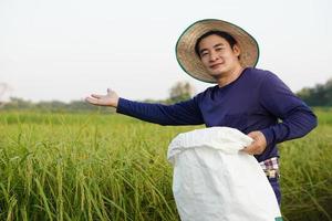 apuesto agricultor asiático está en el campo de arroz, usa sombrero, sostiene un saco blanco de fertilizante orgánico, se siente seguro. concepto, agricultor satisfecho con el producto para la agricultura. foto